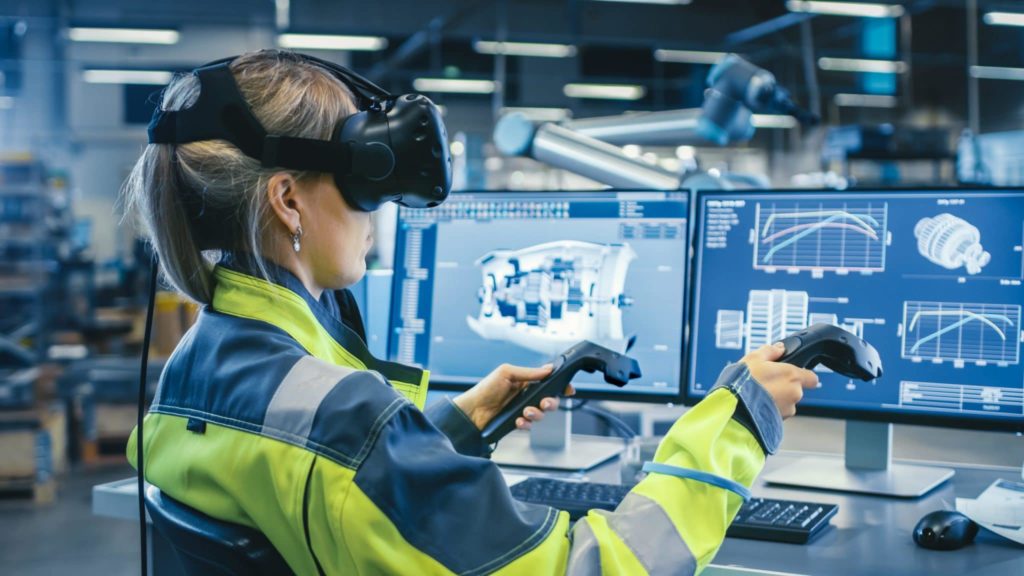 A person wearing virtual reality gear in front of computer screens in a factory.